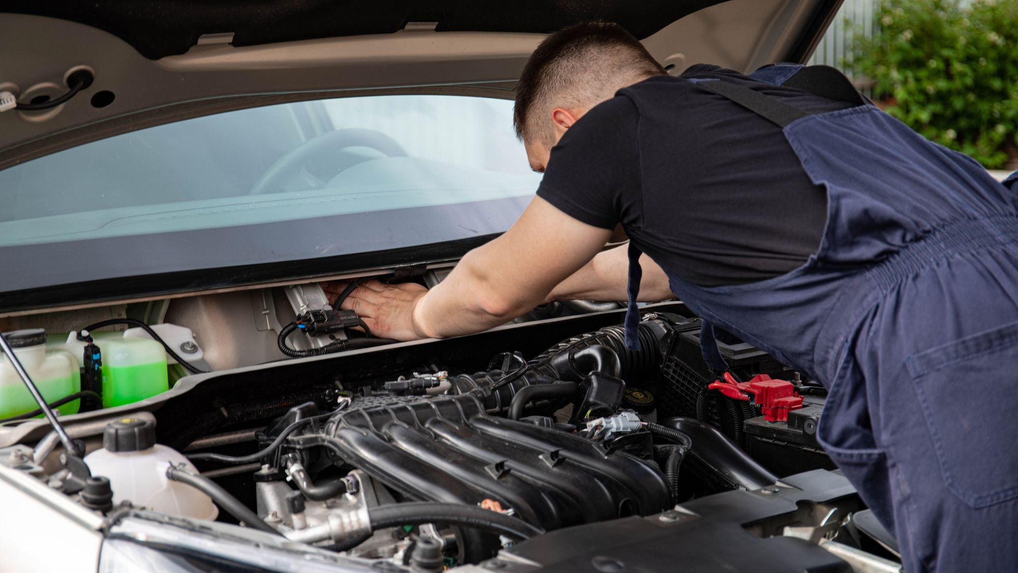 An auto electrician leaning into the engine bay of a car parked outdoors.